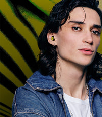 A close-up portrait of a person with dark hair wearing a bright yellow Amnesia Ibiza x Loop Experience  in-ear earbud, styled with a denim jacket over a white t-shirt against a yellow and black striped background.
