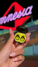 A hand with manicured nails holds an open black wireless earbud charging case containing Amnesia Ibiza x Loop Experience two bright yellow earbuds. The background features a blurred red and purple neon sign with the word 'Amnesia'.