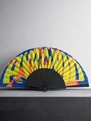A folding hand fan with a black ribbed frame and a bright yellow, blue, and orange abstract patterned canopy, displayed on a dark surface against a light gray wall. The text 'IBIZA DANCE CREW' is printed on the lower left of the canopy.